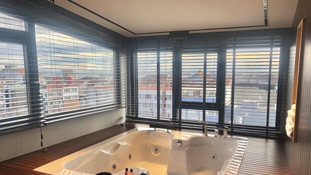 Un baño moderno con una bañera grande, suelo de madera y ventanas amplias con persianas.