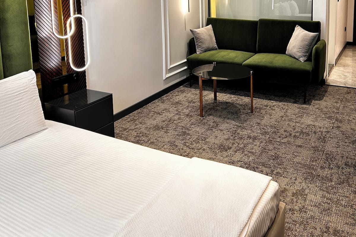 Une chambre d’hôtel moderne avec un lit, un canapé vert, une petite table et une cloison en verre.