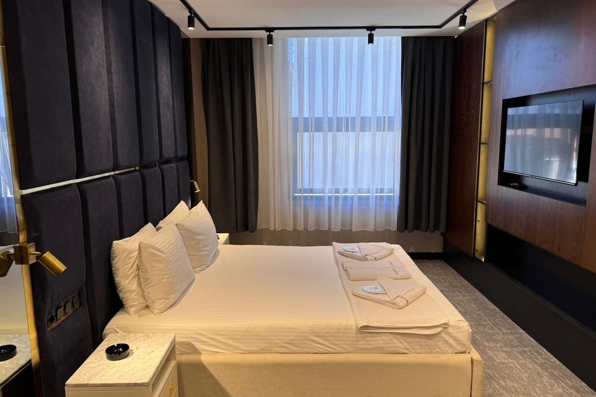 Une chambre d’hôtel avec un grand lit, une table de chevet, un téléviseur fixé au mur et une fenêtre avec des rideaux.