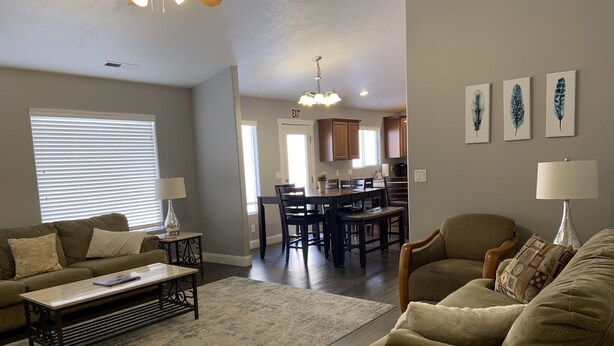 A living room with a sofa, coffee table, and a rug. There is a dining area with a table and chairs. The kitchen is visible in the background with cabinets and a ceiling light. - The Zion Getaway-Located in beautiful Southern Utah (La Verkin)