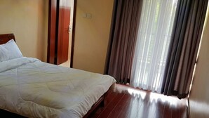 A room with a bed, wooden flooring, a door, and a window with curtains. - Riverland Apartments (Nairobi)