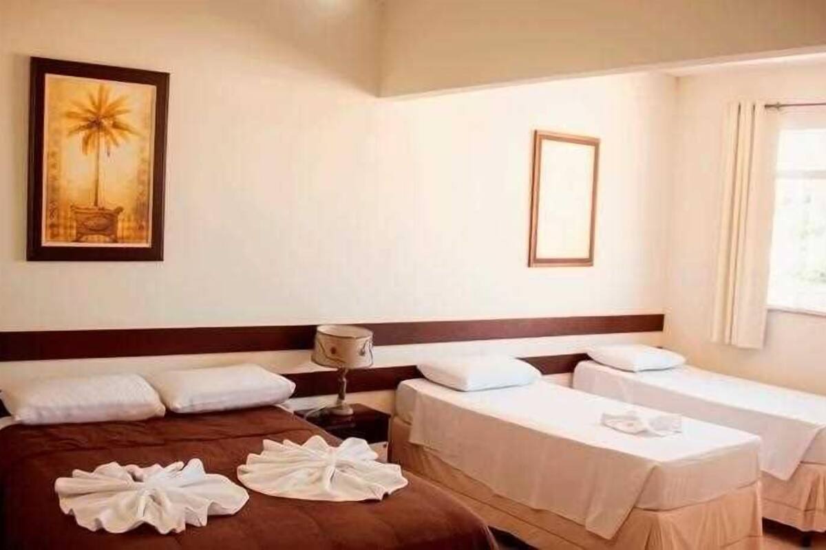 Hotellihuone, jossa on suuri sänky, kaksi pienempää sänkyä, yöpöytä lampun kanssa, seinällä oleva kehystetty kuva ja ikkuna, jossa on verhot.