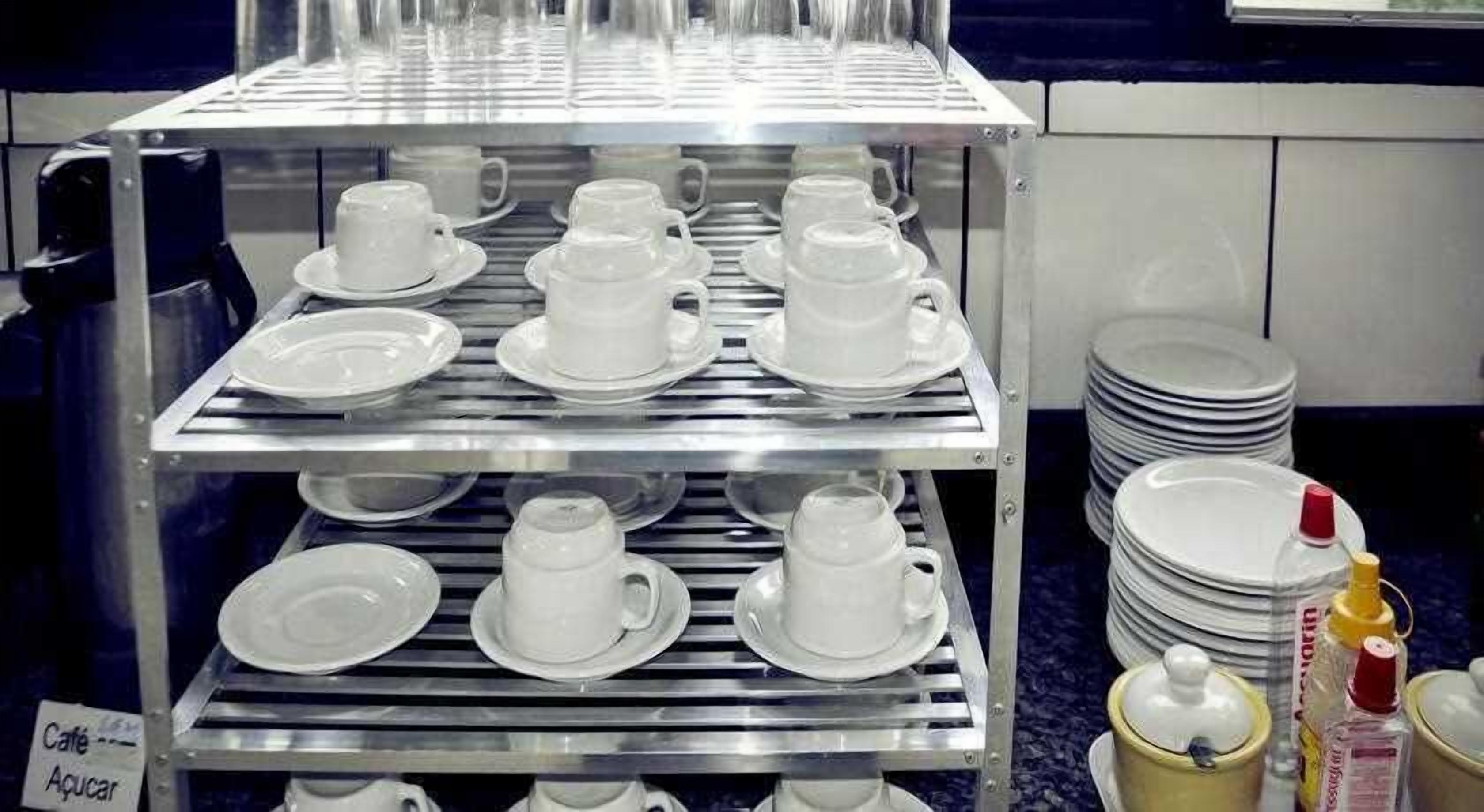 Uma estante de aço inoxidável com várias fileiras de xícaras e pires brancos, pratos empilhados e temperos.
