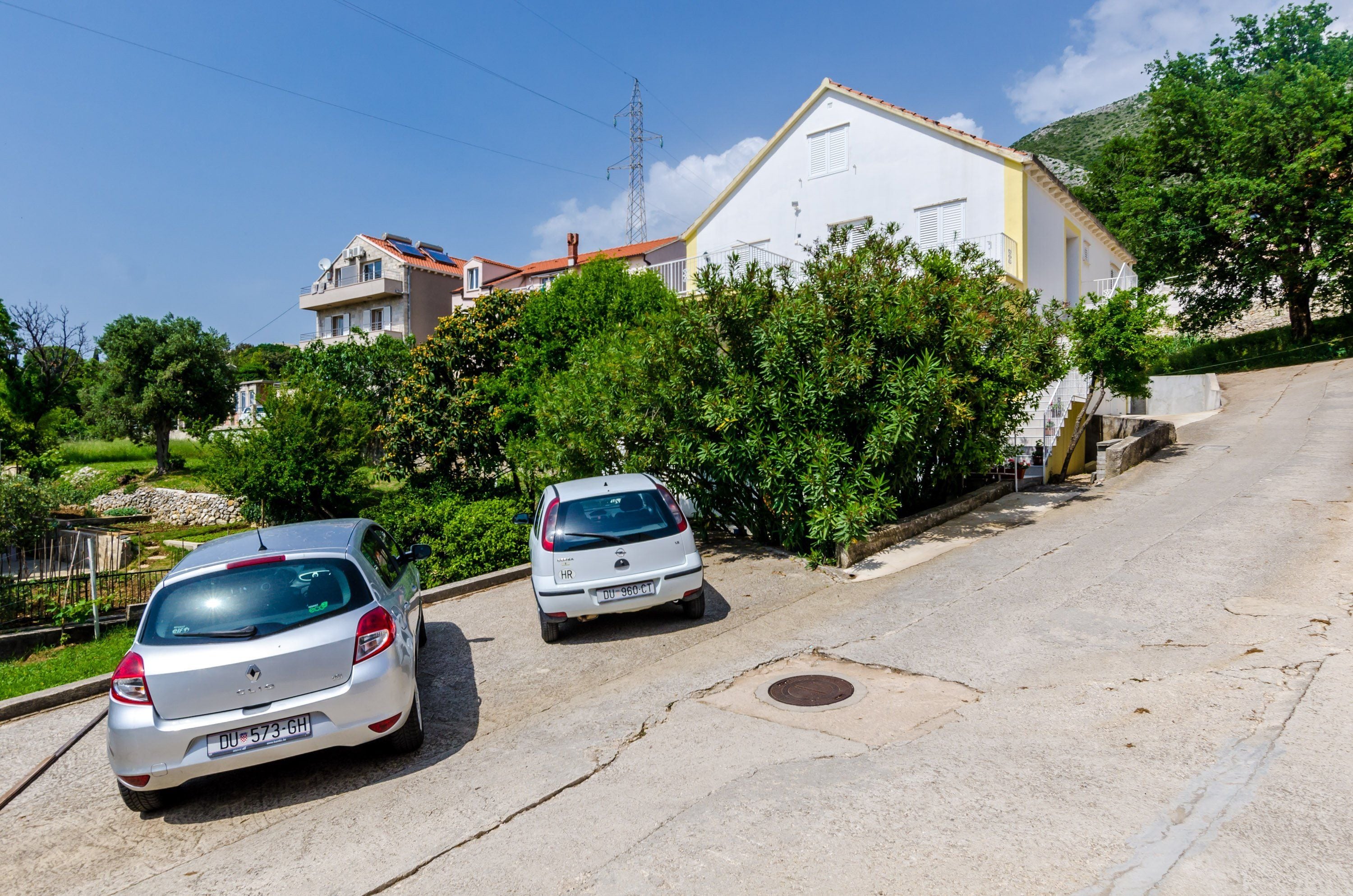 Rua residencial com dois carros estacionados, um prédio branco com telhado vermelho e vegetação exuberante.