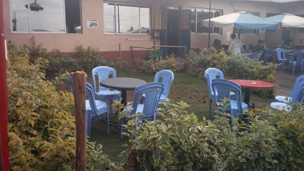 A building with a sign indicating fish availability, outdoor seating with plastic chairs, and a covered area with umbrellas. - Small Villa Gardens (Kabati)