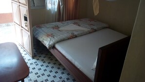 A single bed with a floral bedspread, a wooden headboard, and a mosquito net. There is a small table to the left and a window with curtains. - Small Villa Gardens (Kabati)