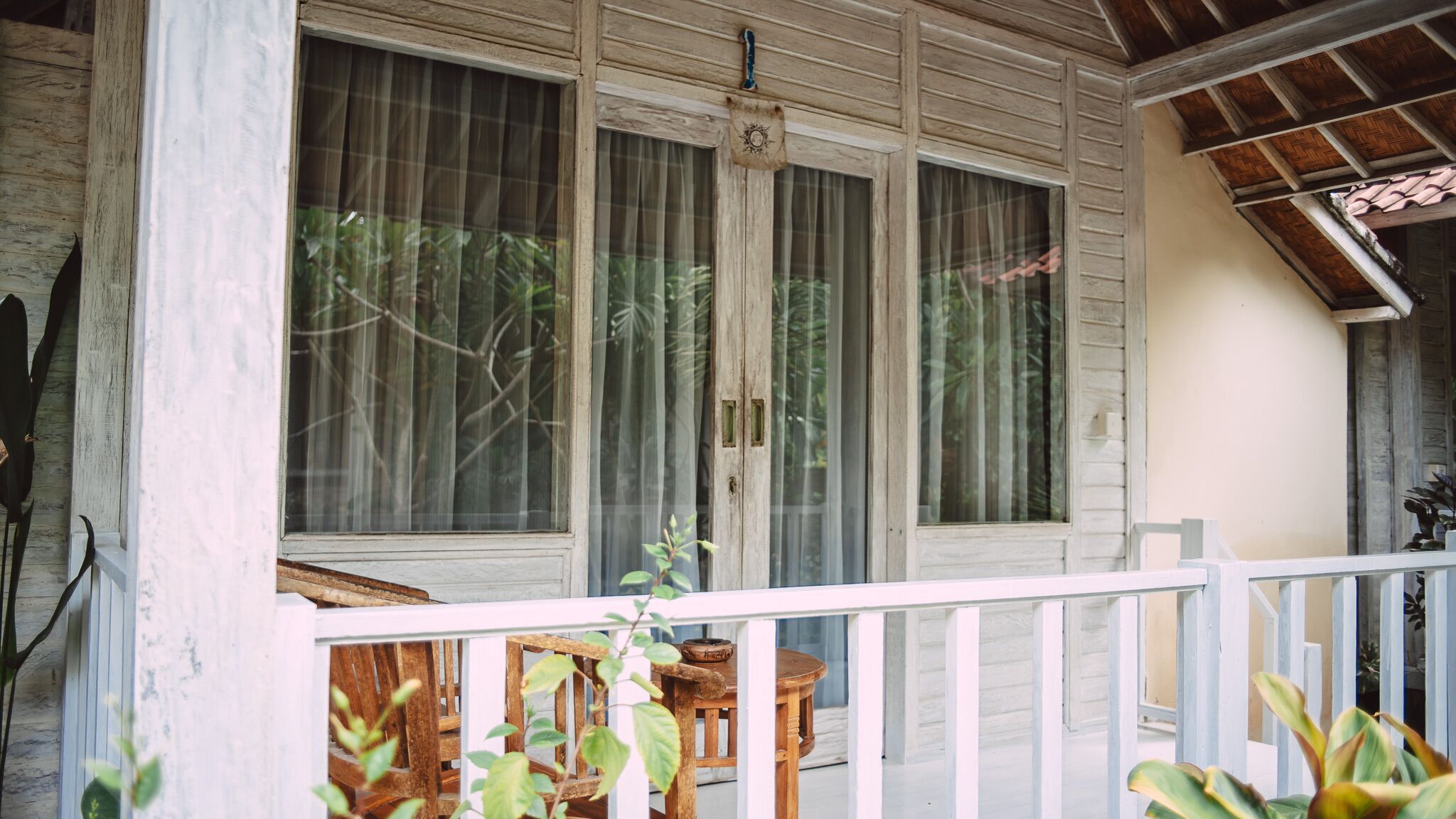 A wooden porch with a white railing, large windows with curtains, and a small table.