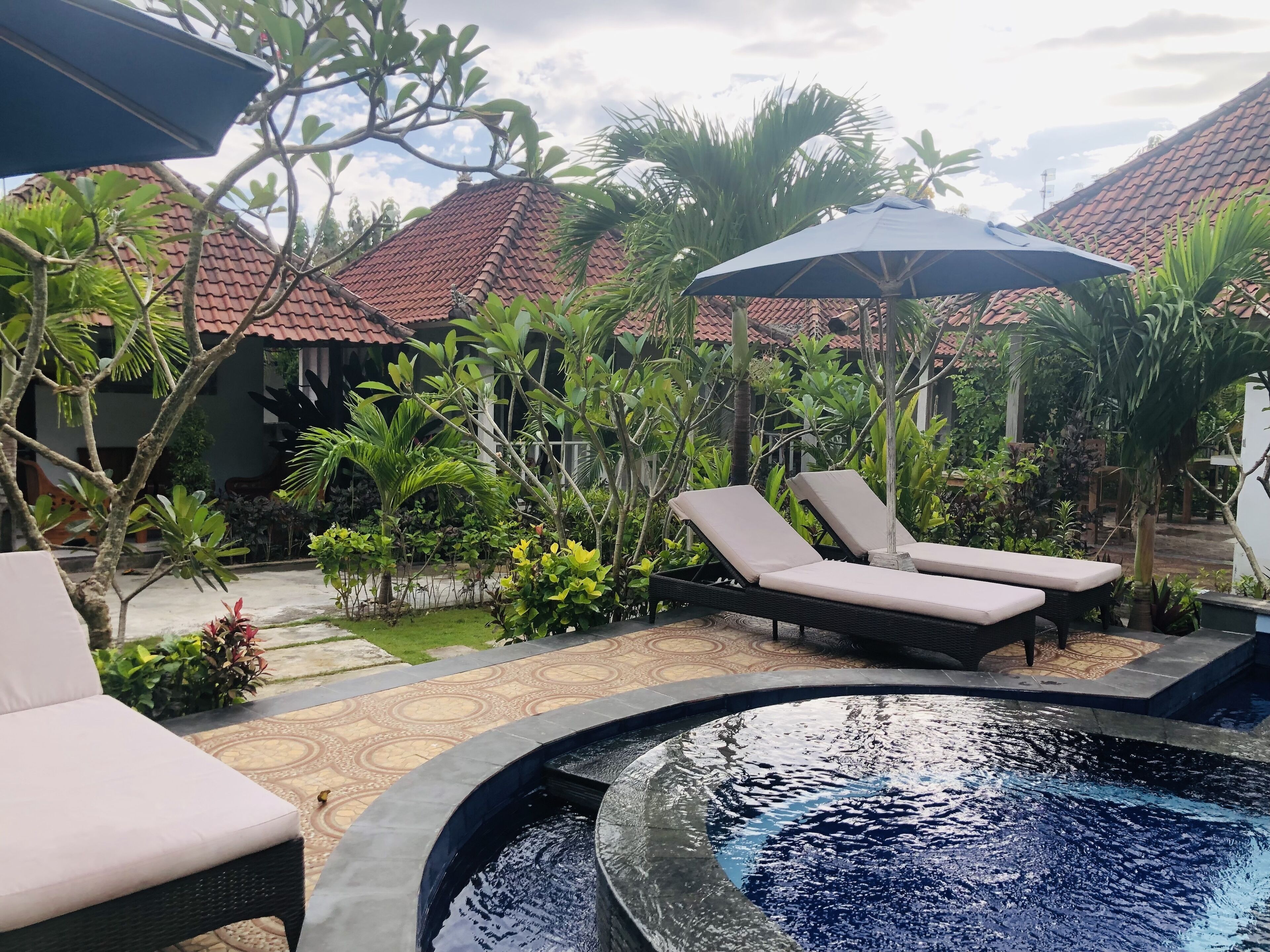 A pool area with lounge chairs and umbrellas, surrounded by tropical plants and a tiled roof structure.
