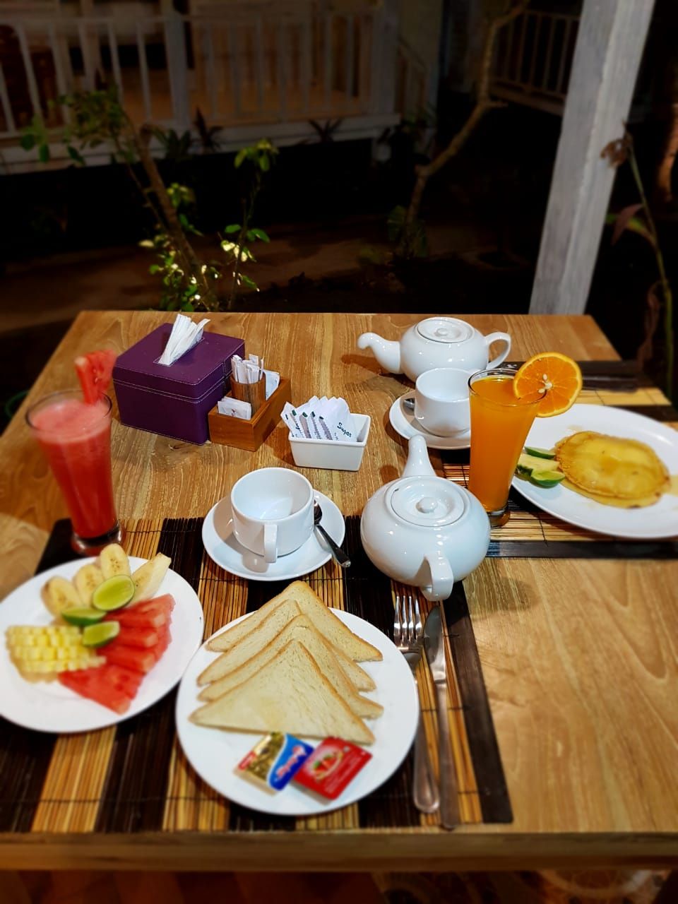 A breakfast setup with a teapot, cups, a glass of orange juice, and plates of toast and fruit.