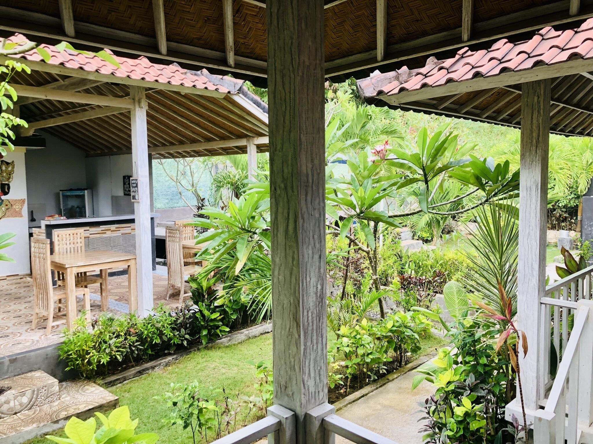 A covered outdoor area with a wooden table and chairs, surrounded by lush greenery and a tiled roof.