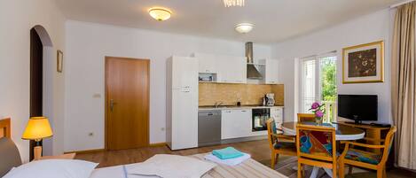 A hotel room with a bed, a desk with a TV, a dining area with chairs, a kitchen with white cabinets, and a door leading to another room.