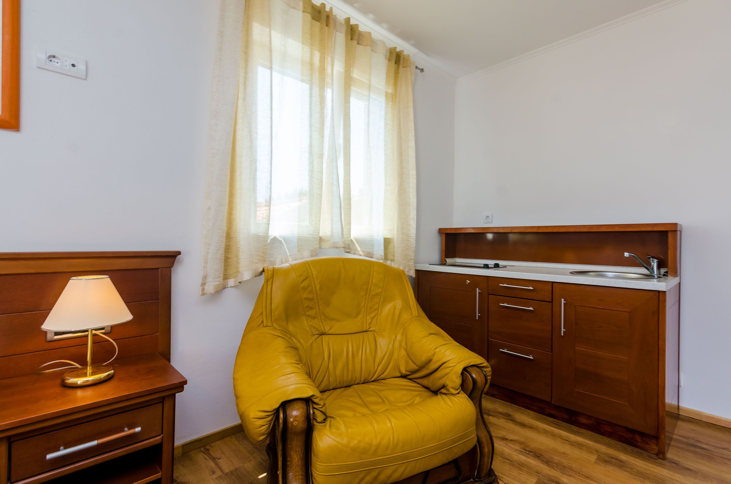 A compact room with a wooden chair, a small table with a lamp, a kitchenette with a sink, and a window with curtains.