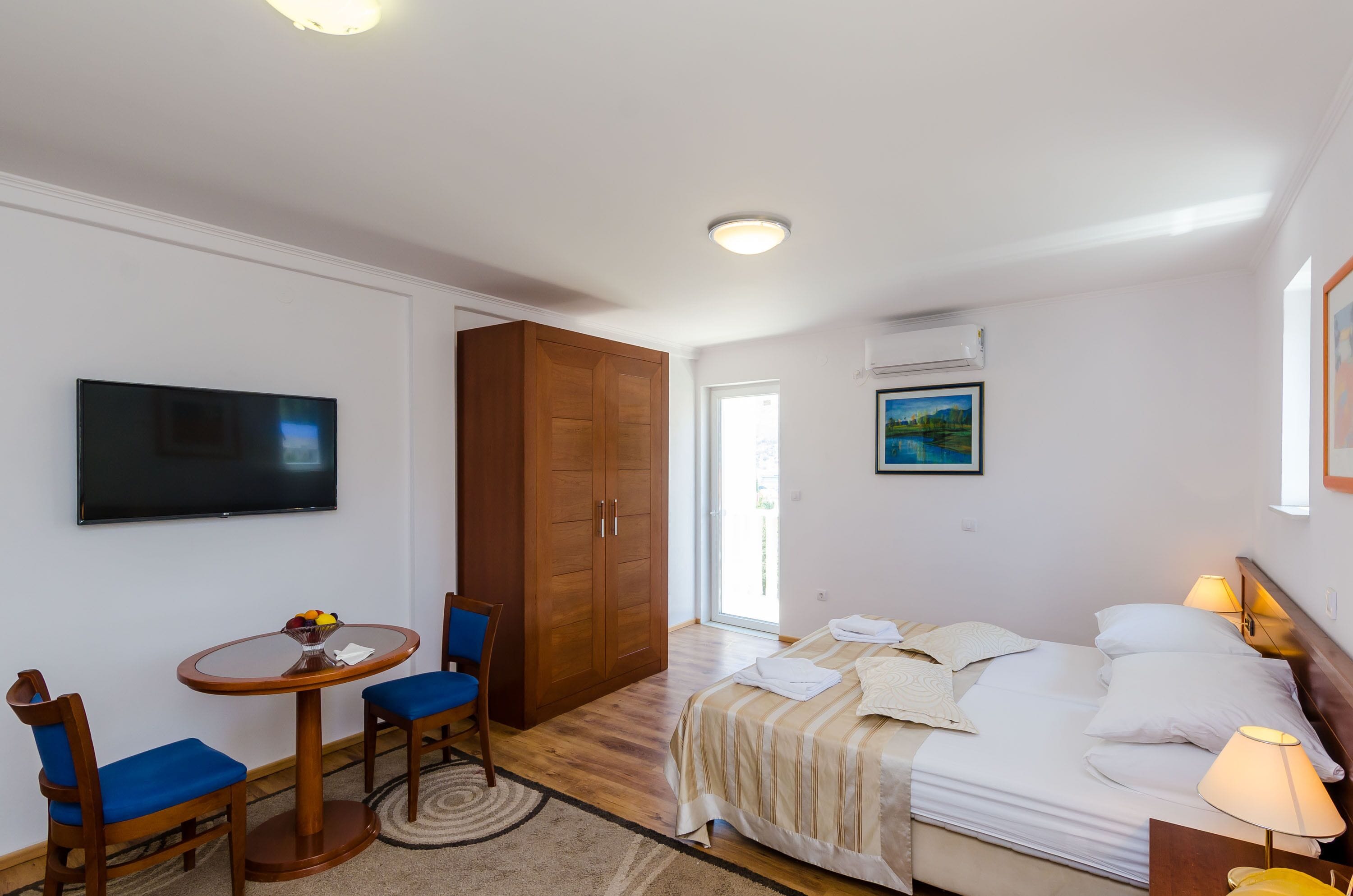 A hotel room with a bed, a TV, a wooden wardrobe, a round table with chairs, and a window with a view.
