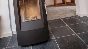 A modern fireplace with a black exterior and a glass front, set in a room with dark tiled flooring and a wooden shelf in the background. - Winzerhaus am Eckbach (Bissersheim)