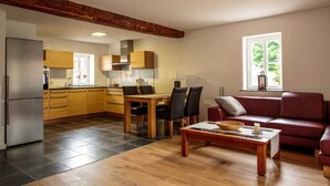 A modern kitchen with wooden cabinets, a dining table with chairs, and a red leather sofa in a living area with a large window. - Winzerhaus am Eckbach (Bissersheim)