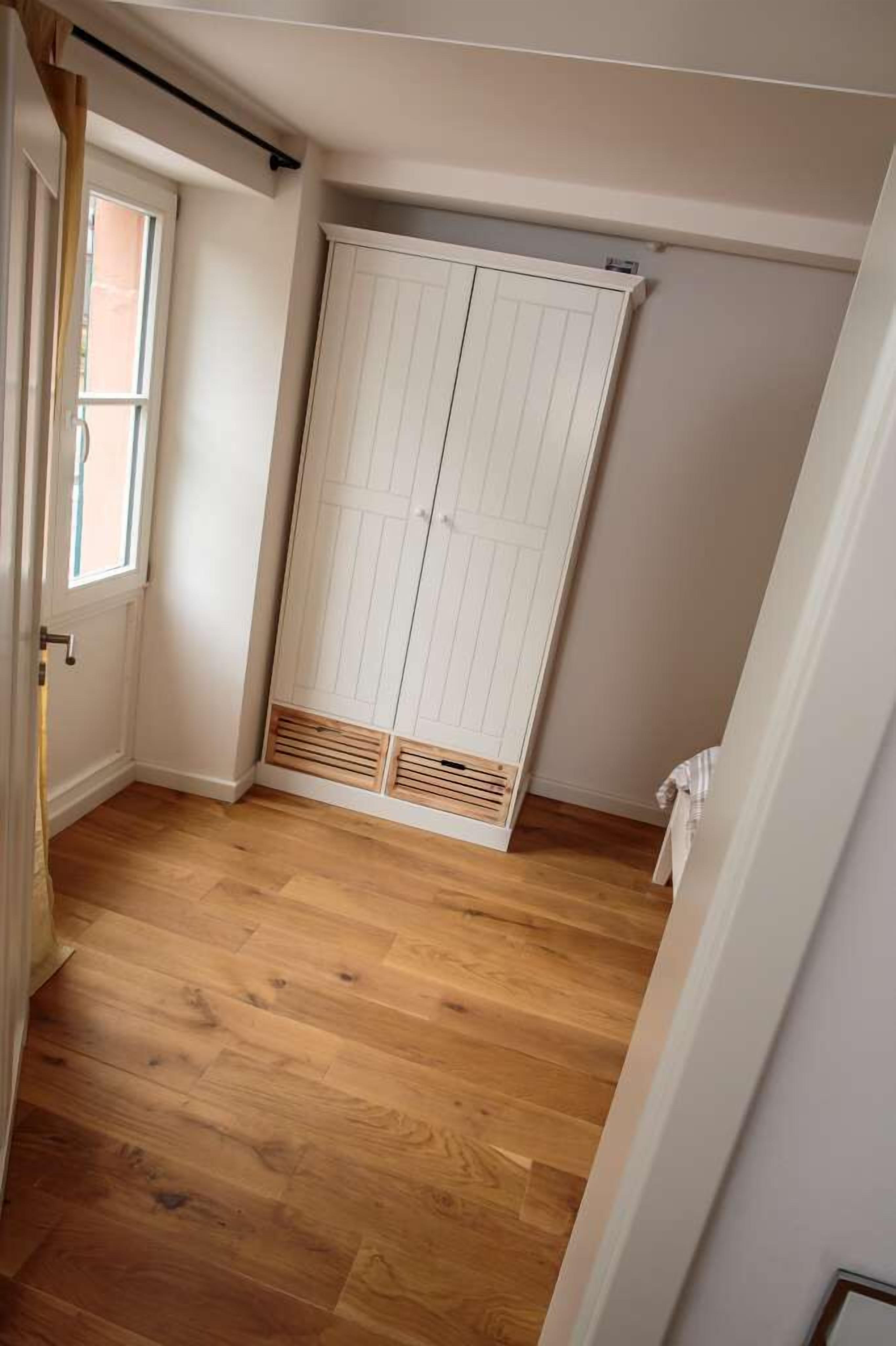 Una habitación con suelo de madera, un armario blanco con rejilla de ventilación y una ventana.