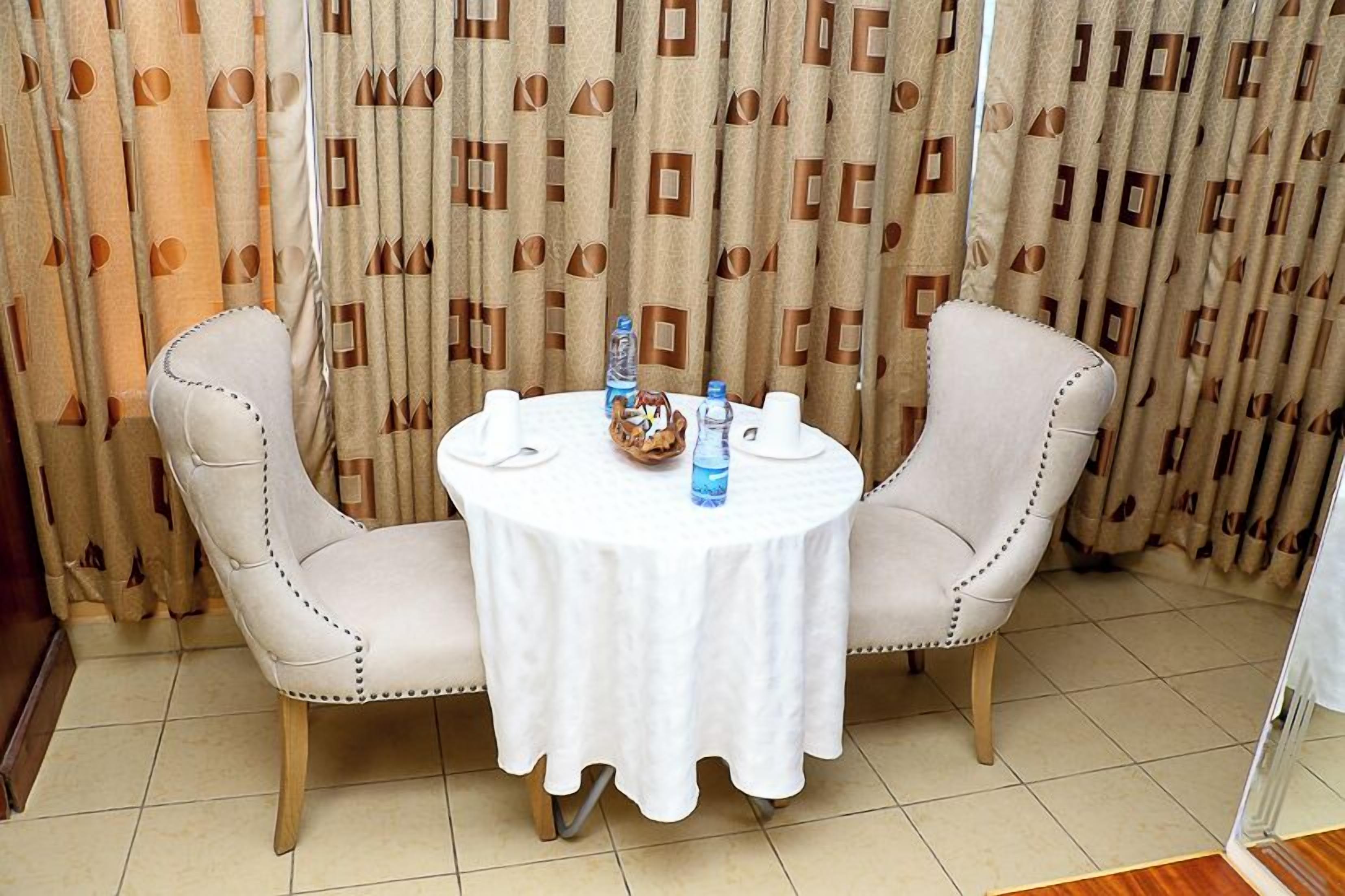 Una habitación con una mesa redonda para dos personas, dos sillas tapizadas, una botella de agua, un bol y dos tazas blancas.