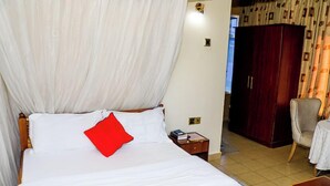 Una cama bien tendida con sábanas blancas y una almohada roja, una mesita de noche de madera con un libro, una silla y un armario en una habitación con paredes beige y cortinas.
