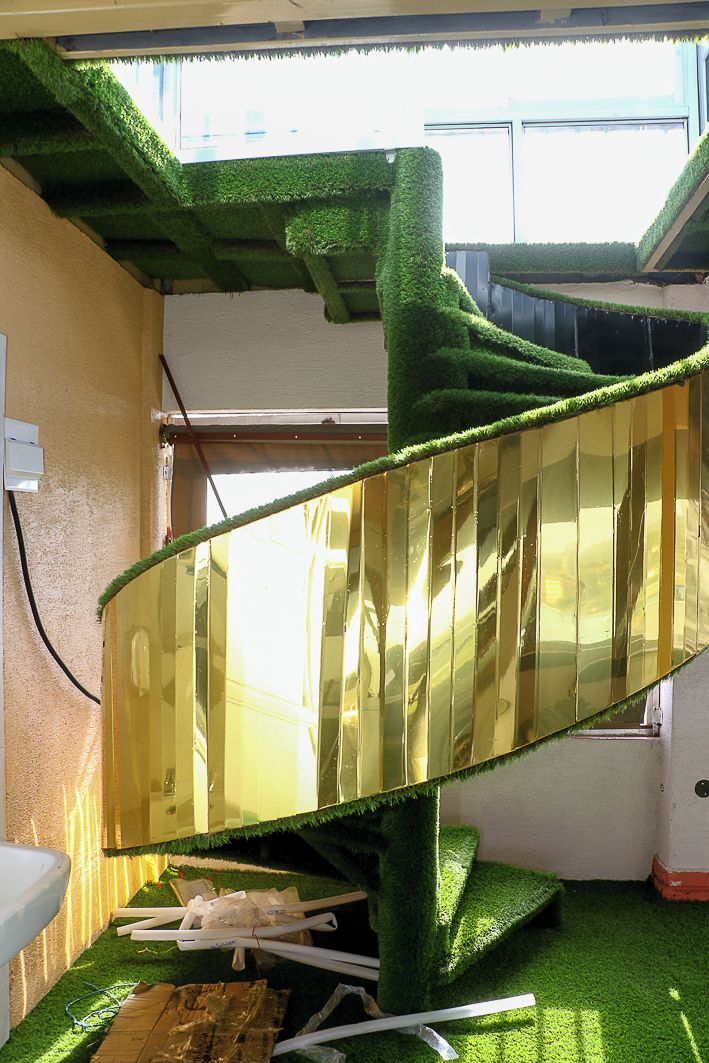 A room with a green artificial grass floor, a large mirror reflecting the room, and a window with blinds.