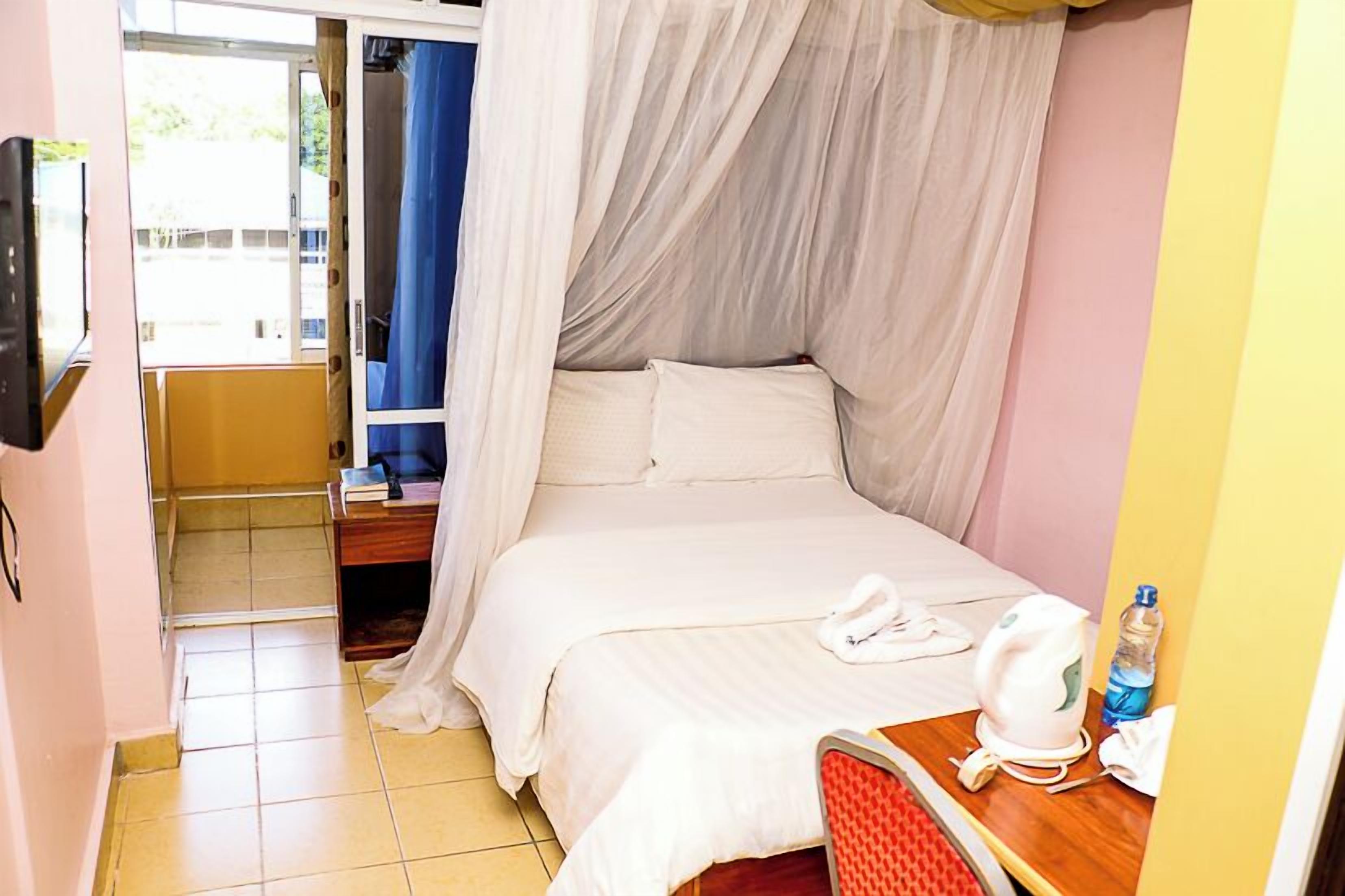 A single bed with a canopy, a bedside table with a kettle and a bottle, and a chair.