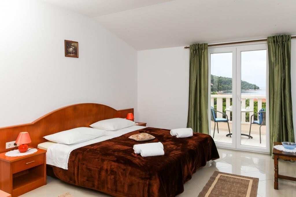 A hotel room with a large bed, a nightstand, a lamp, and a view of the outdoors through a sliding glass door.