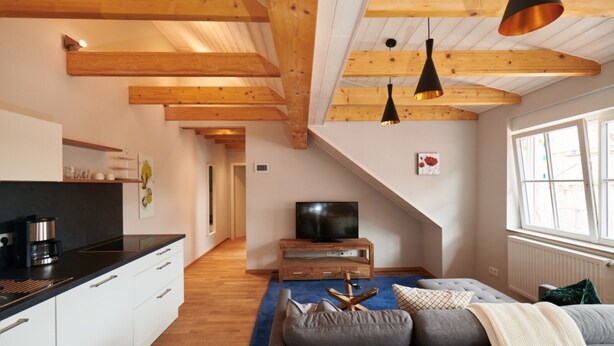 Un espacio habitable moderno con cocina, sofá y televisor en una habitación estilo loft con vigas de madera a la vista y un ventanal grande.