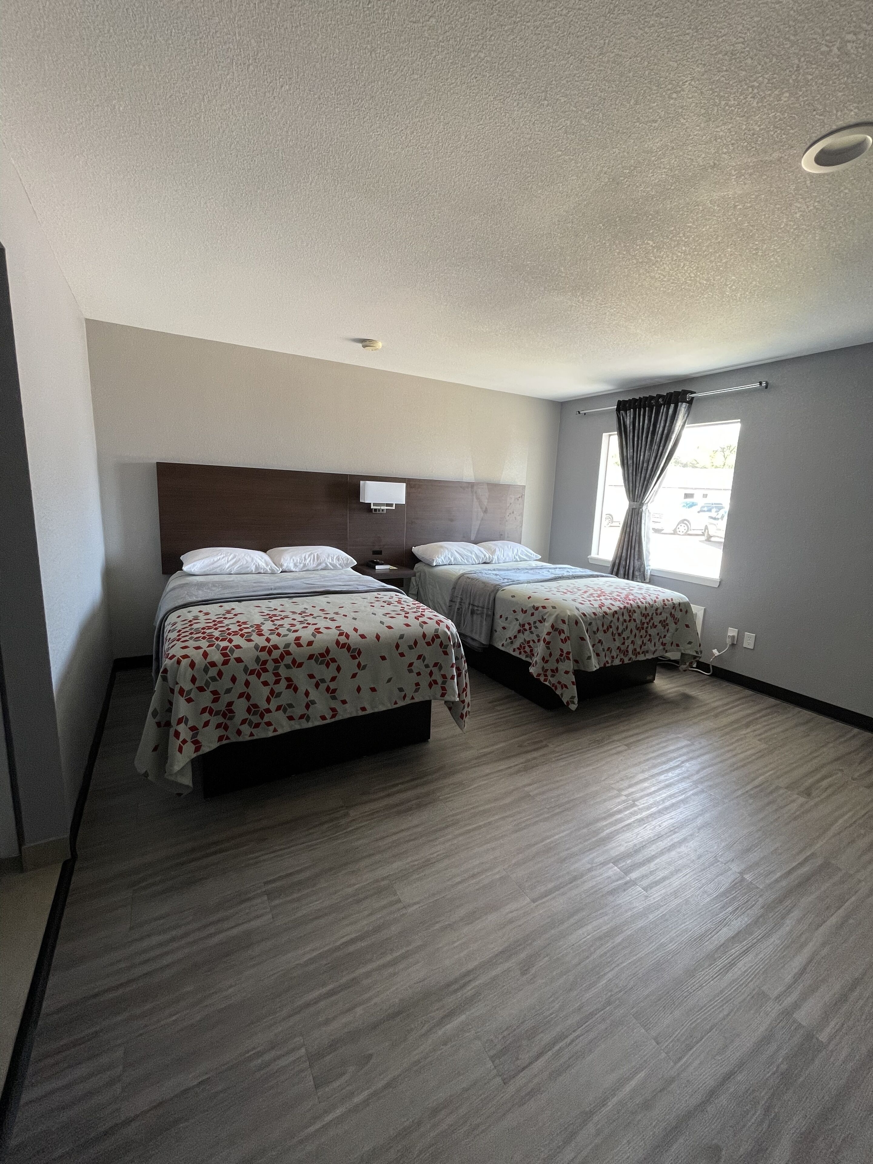 Une chambre d’hôtel avec deux lits, un plancher en bois, une tête de lit, une fenêtre avec des rideaux et un luminaire fixé au mur.