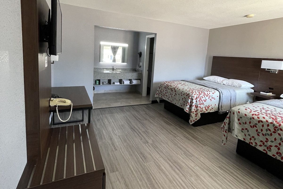 A hotel room with two beds, a desk with a telephone, a television mounted on the wall, and a bathroom visible through an open door.