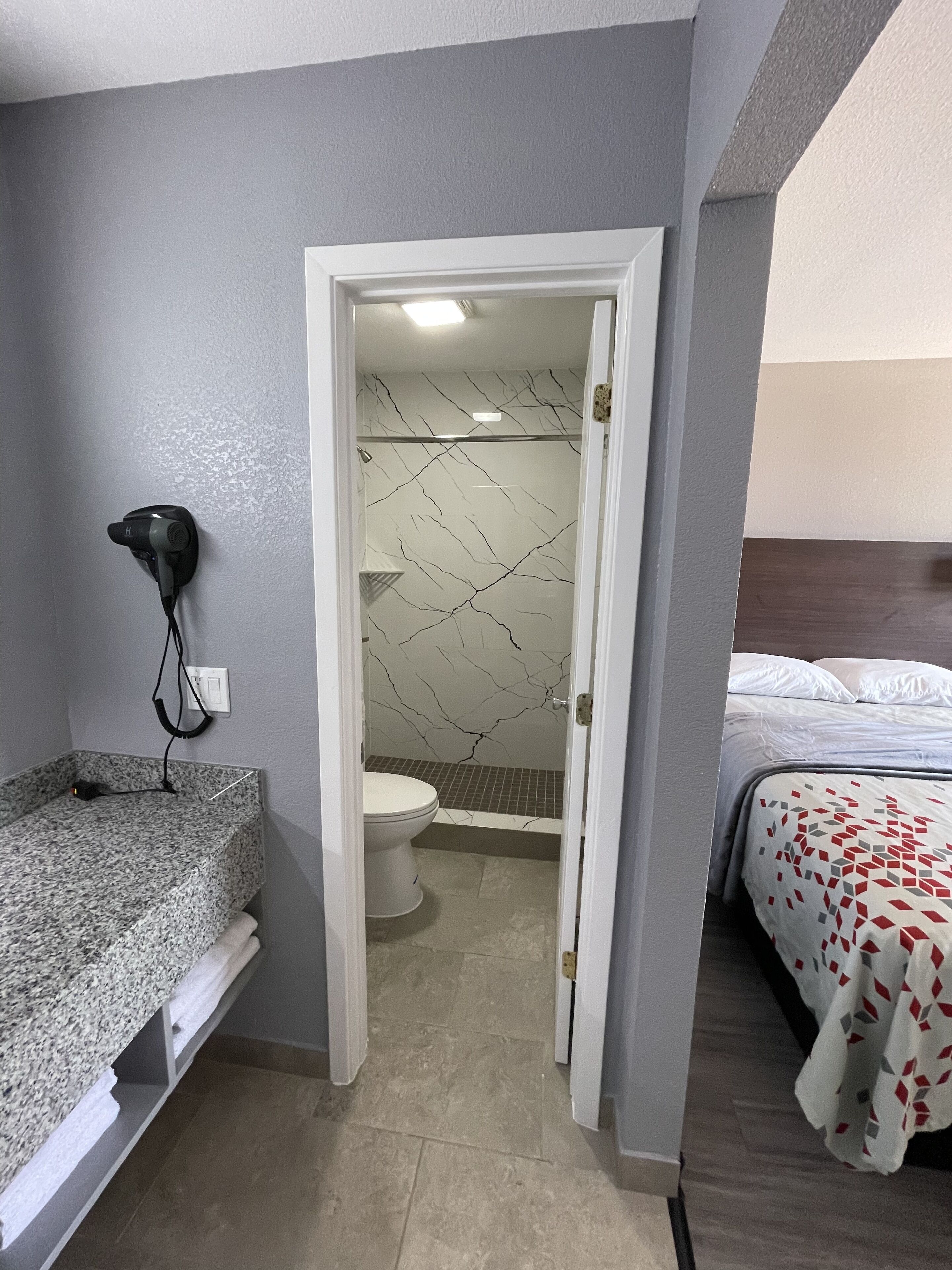 A compact hotel room with a shower, toilet, and a bed with a patterned bedspread.
