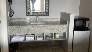 A bathroom with a granite countertop, a sink, a mirror, a hairdryer, a trash bin, and a refrigerator.