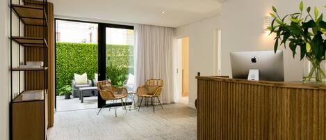 A modern hotel room with a wooden reception desk, a laptop, and a potted plant. There is a seating area with wicker chairs and a view of a green hedge outside.