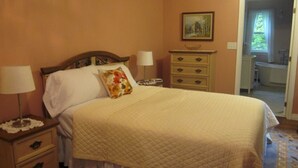 A neatly made bed with a quilted cover, a bedside table with a lamp, a dresser with drawers, and a painting on the wall. - Chestnut Lane Bed & Breakfast - The Rose Room (New Glasgow)