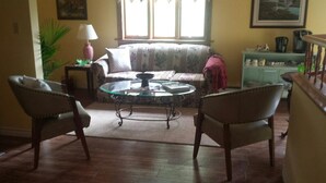 A living room with a sofa, two armchairs, a glass coffee table, a lamp, a plant, and a window with curtains. - Chestnut Lane Bed & Breakfast - The Rose Room (New Glasgow)