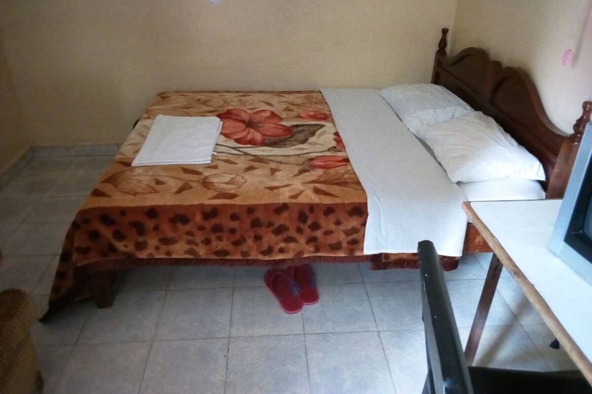 A room with a bed, a wooden headboard, a white pillow, a floral bedspread, and a pair of red slippers on the floor.