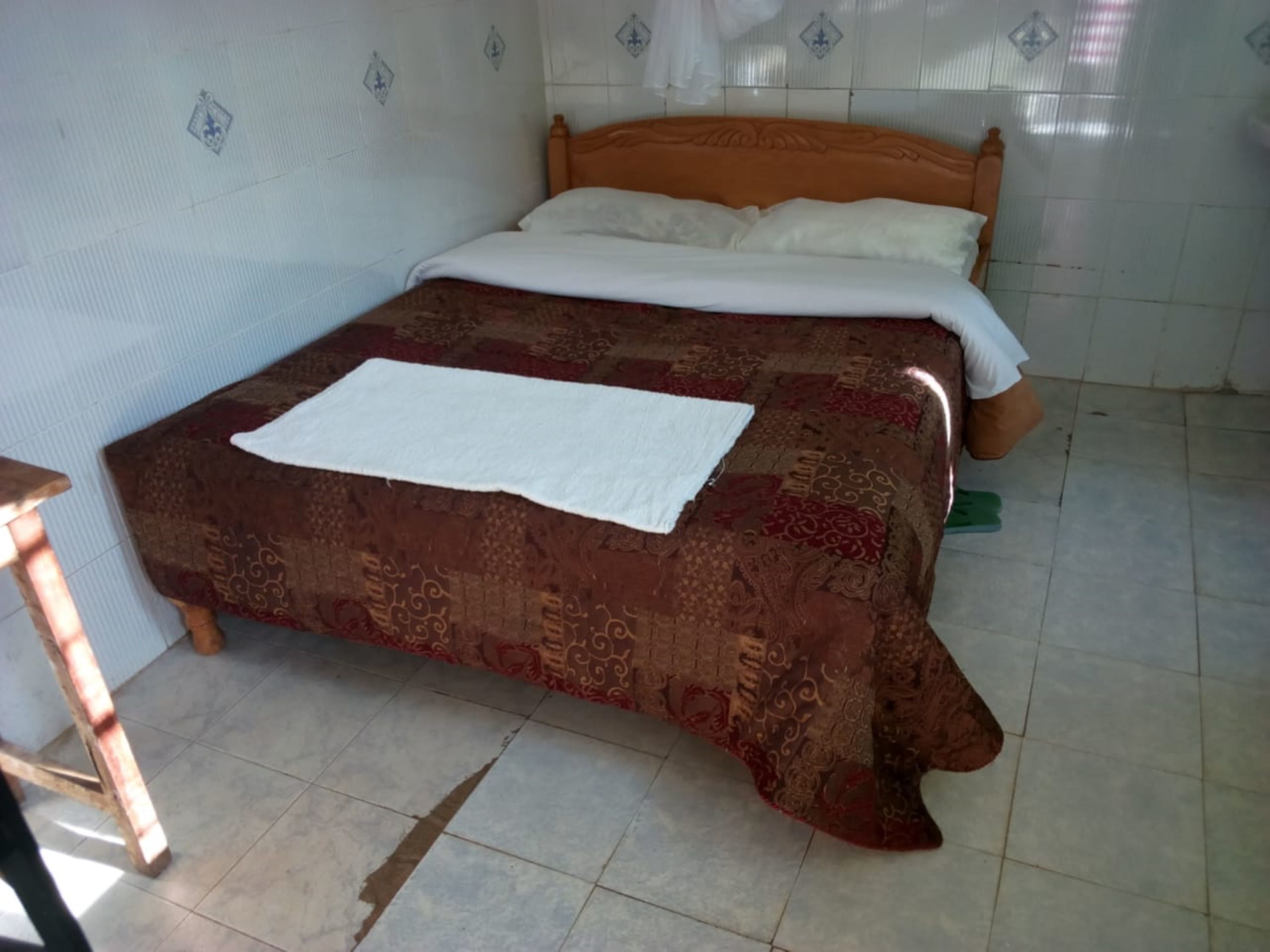 A bed with a patterned bedspread and white pillows, a wooden headboard, and a small wooden stool to the left.