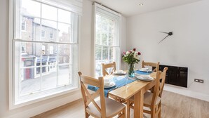 Comedor con mesa de madera dispuesta para dos personas, ventanas amplias y chimenea.
