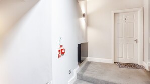 Un pasillo con una puerta blanca marcada con el número 2, un detector de incendios y una alfombra de entrada con un mensaje.