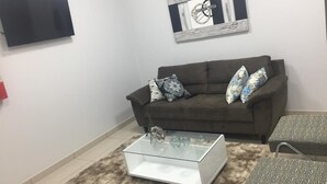 Una sala de estar con un sofá, una mesa de centro de vidrio, un reposapiés estampado, un televisor montado y un espejo decorativo.