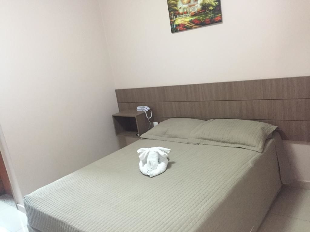 Een net opgemaakt bed met een gevouwen handdoek, een nachtkastje met een lamp en een aan de muur hangend schilderij.