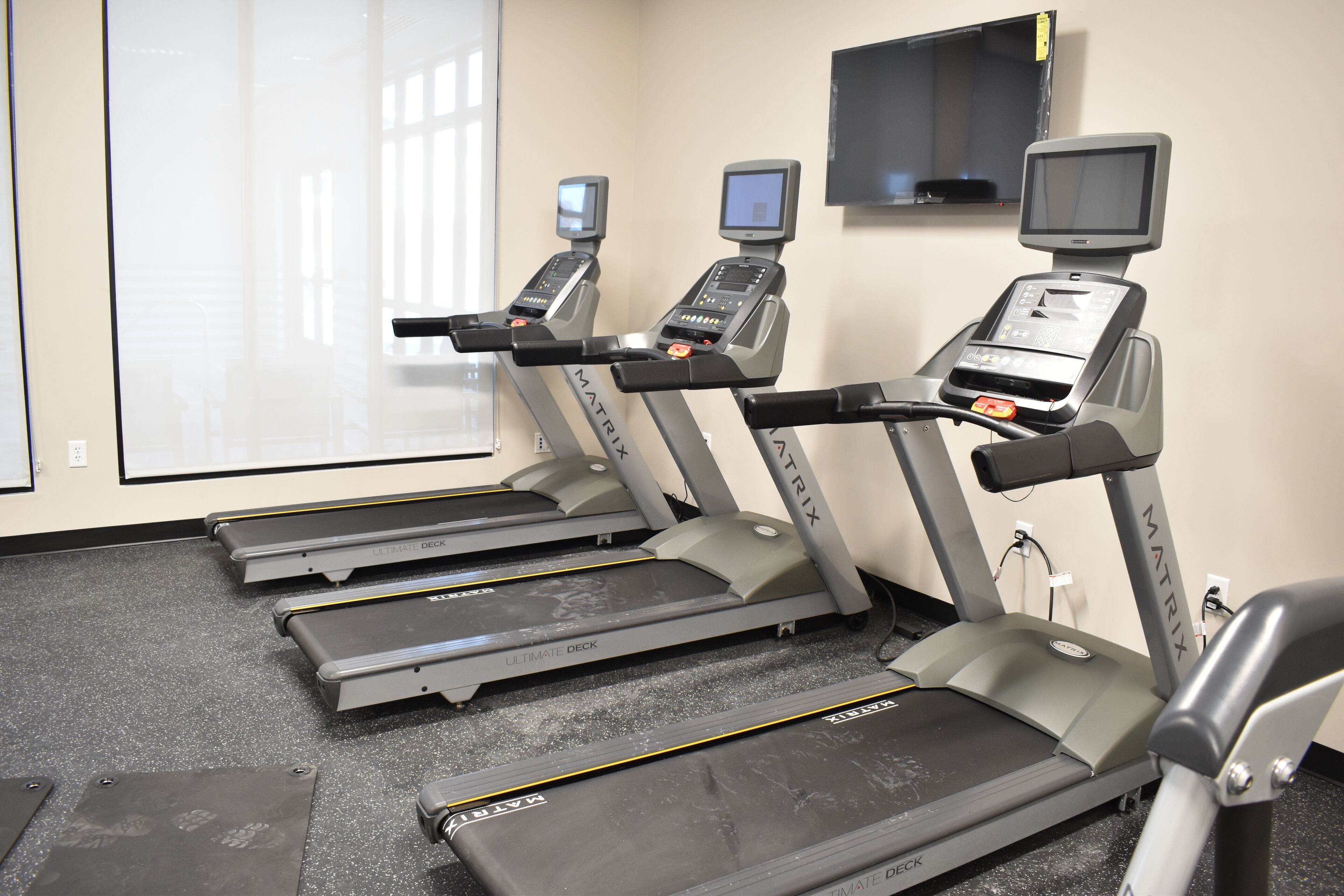 A gym with treadmills, each equipped with a digital display, in a room with a large window and a mounted TV.