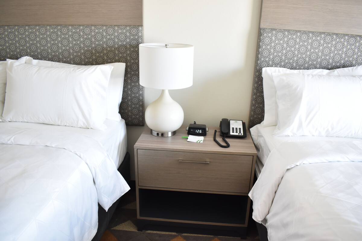 A hotel room with two beds, a bedside table with a lamp, a phone, and a nightstand drawer.