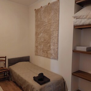 A small, neatly arranged bedroom with a single bed, a chair, a shelf with boxes, and a textured wall hanging.