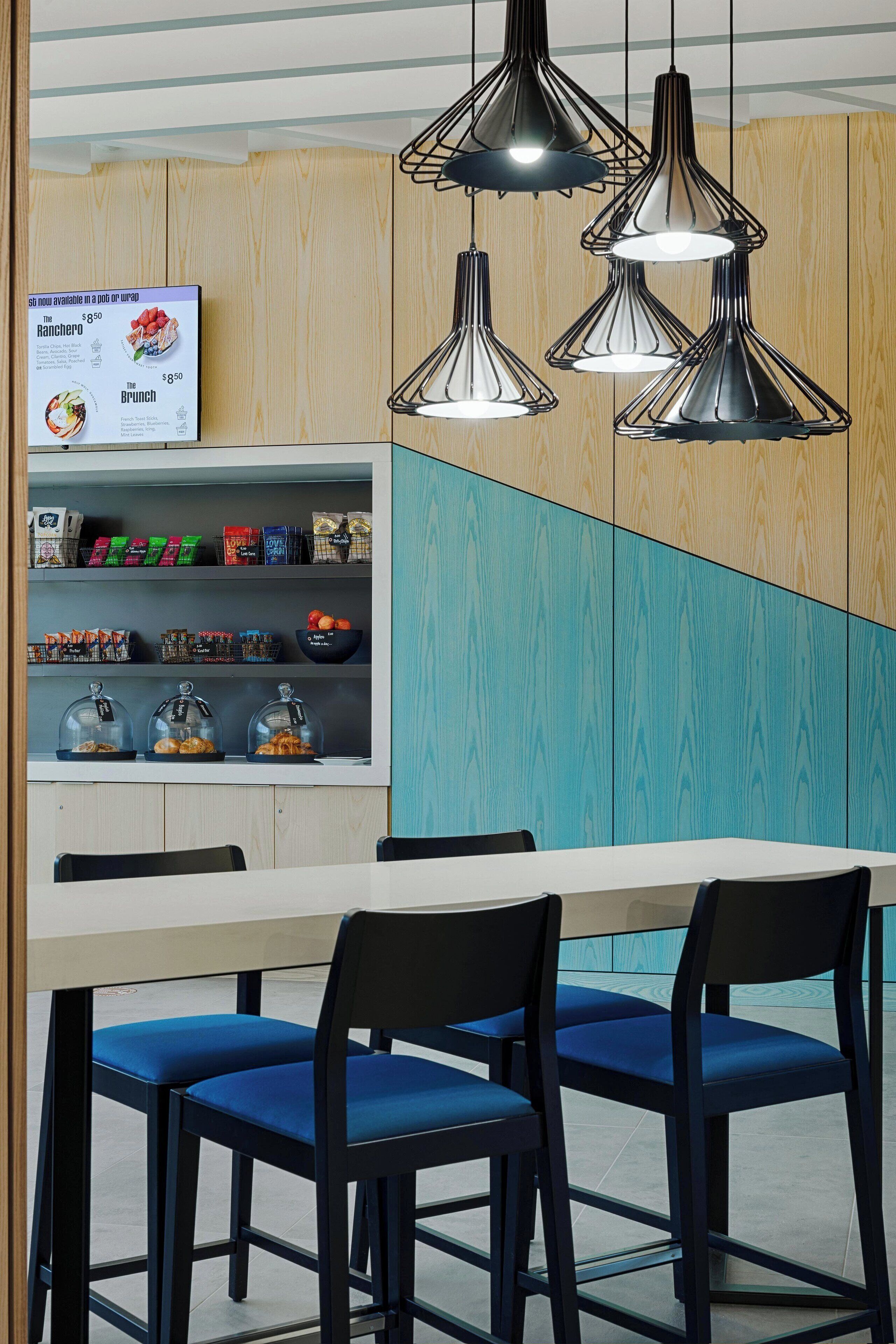 A modern dining area with a wooden and teal color scheme, featuring a white table with blue chairs, a glass display case with snacks, and hanging pendant lights.