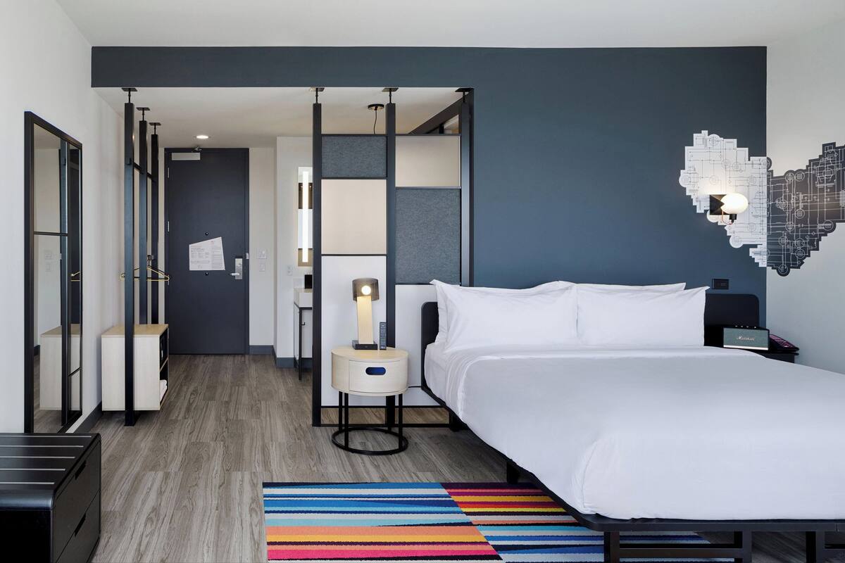A modern hotel room with a large bed, a nightstand with a lamp, a desk with a chair, and a colorful striped rug.