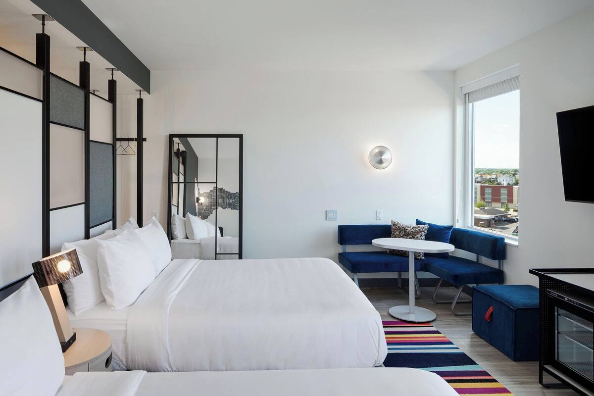 A modern hotel room with a large bed, a seating area with a blue sofa and a small table, a large window with a city view, and a flat-screen TV.