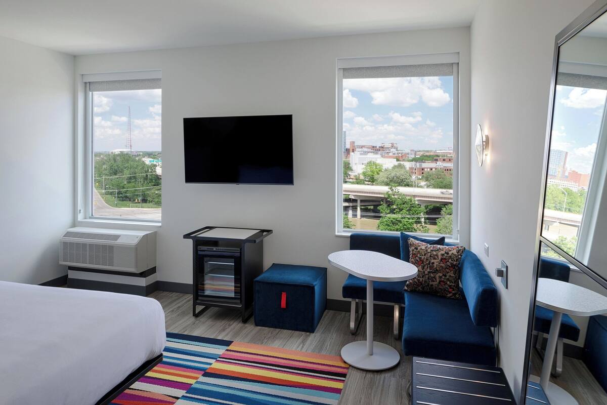 A modern hotel room with a large bed, a TV mounted on the wall, a small table, a sofa, and a colorful striped rug.