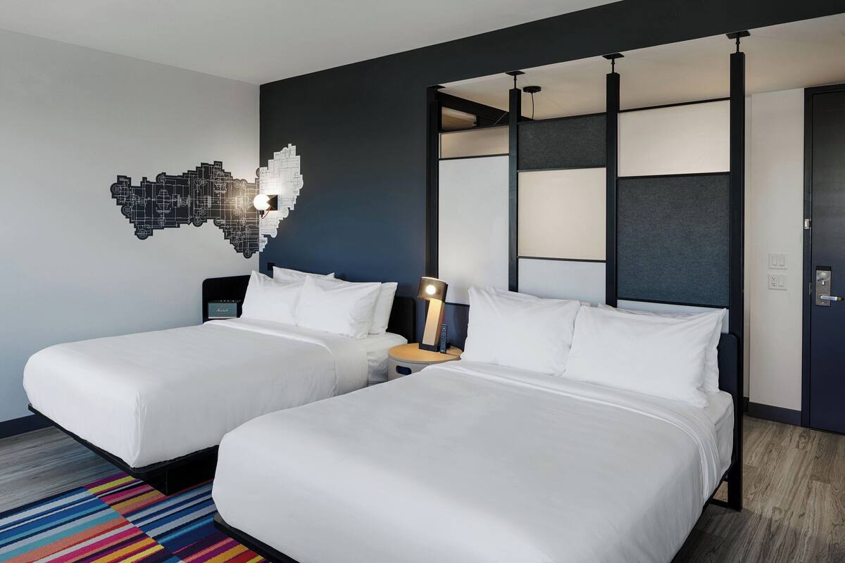 A hotel room with two beds, a modern wall art piece, a nightstand with a lamp, and a colorful striped rug.