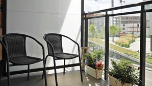 Un balcón con dos sillas de mimbre, una planta en maceta y vistas a una obra en construcción a través de amplias ventanas.