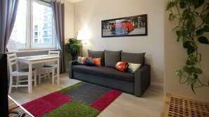 Un moderno salón con un sofá gris, una mesa de comedor con sillas, un ventanal con cortinas, una lámpara de pie, un cuadro colgado en la pared de un coche y una planta en maceta.