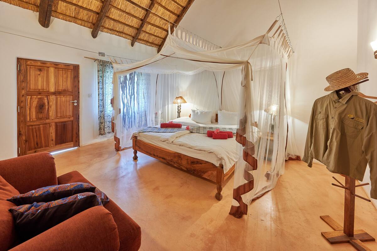 A bedroom with a wooden bed, a canopy, a mannequin dressed in a hat and shirt, and a red chair.